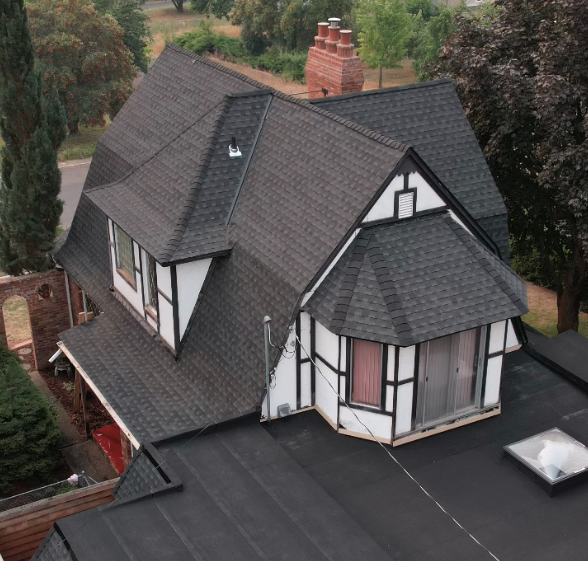 european style home with black shingle roof and brick chimney and a black flat roof