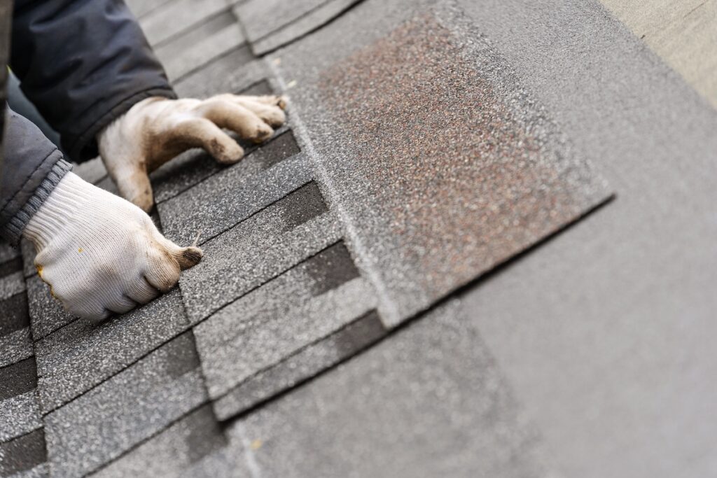 Close up and real photo of skilled workman in special protective work wear installing asphalt or bitumen tile on top of the roof 
